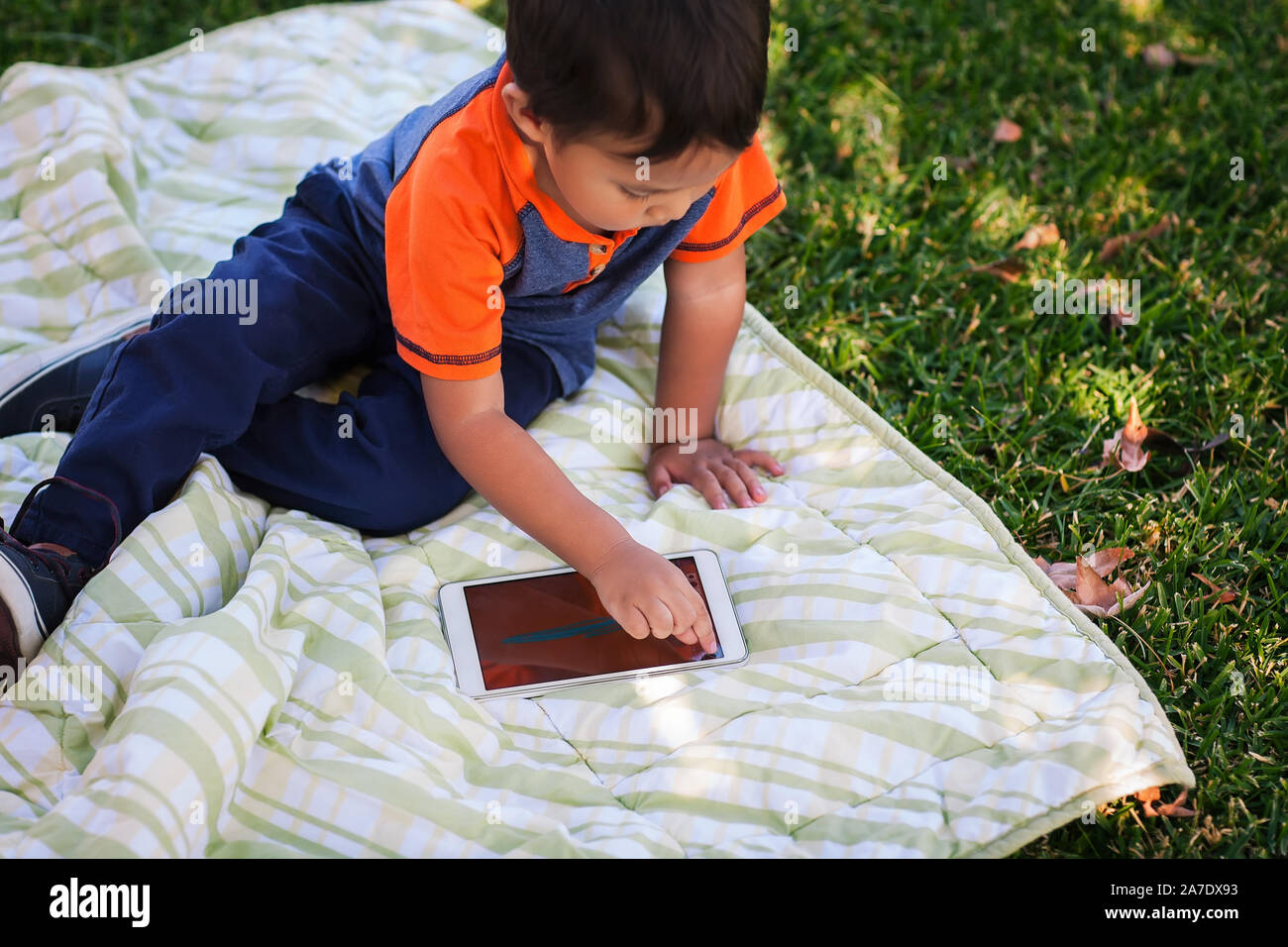 A three year old boy sitting on a picnic blanket and playing on a wifi tablet while resting at a park. Stock Photo