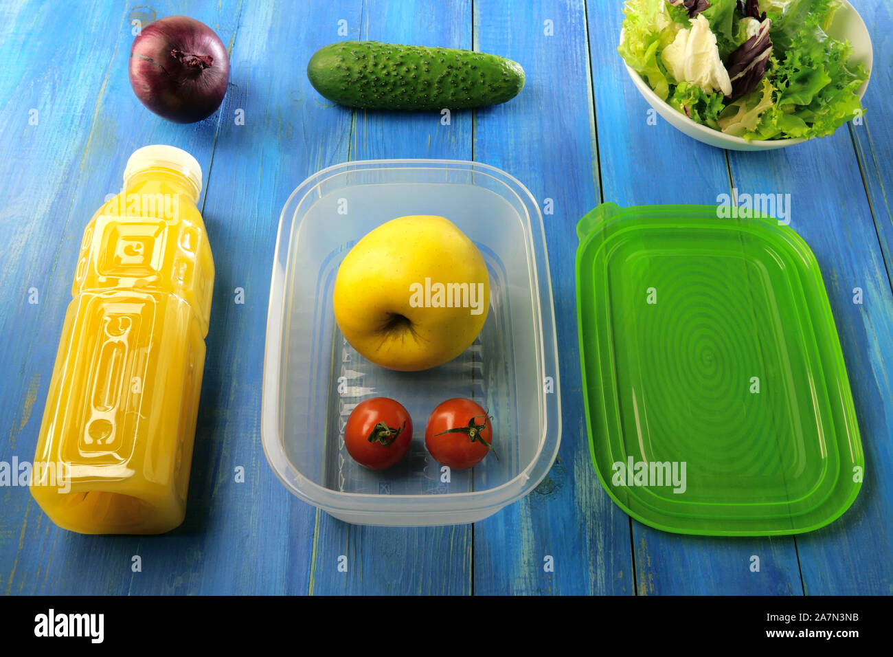 Plastic lunch box with ripe apple, cherry tomatoes and other vegetables on blue wooden table. Nearby is plastic bottle of orange juice and green cap. Stock Photo