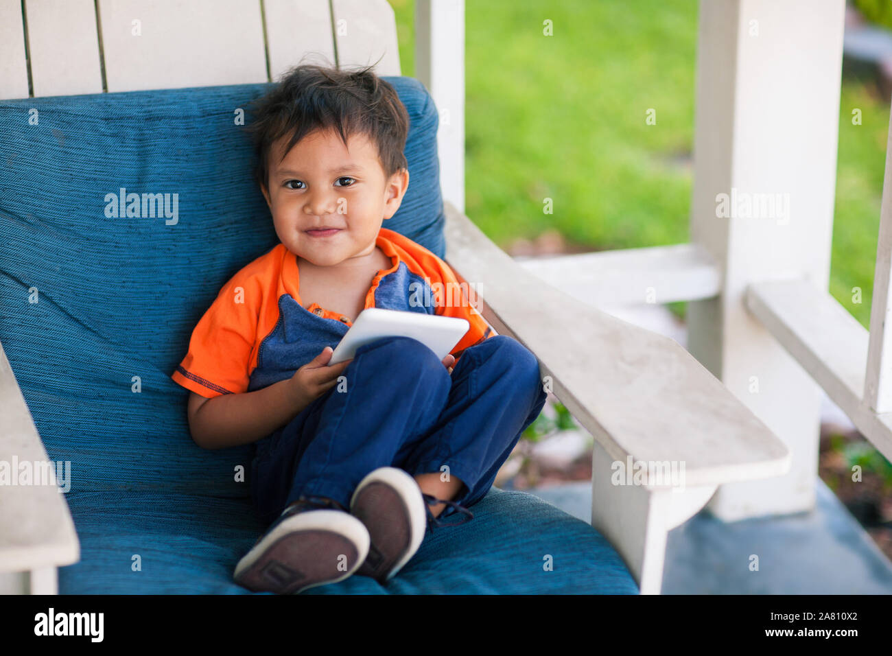 A happy young boy playing educational apps on a white mobile device while sitting in the front porch of his house. Stock Photo