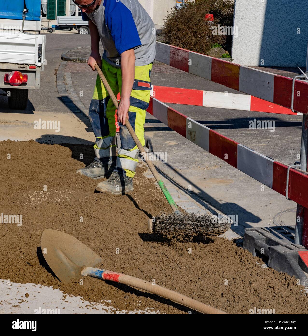 Construction worker with sunglasses and broom scrubbing sand in construction site in front of a construction vehicle and trailer. Construction worker, Stock Photo