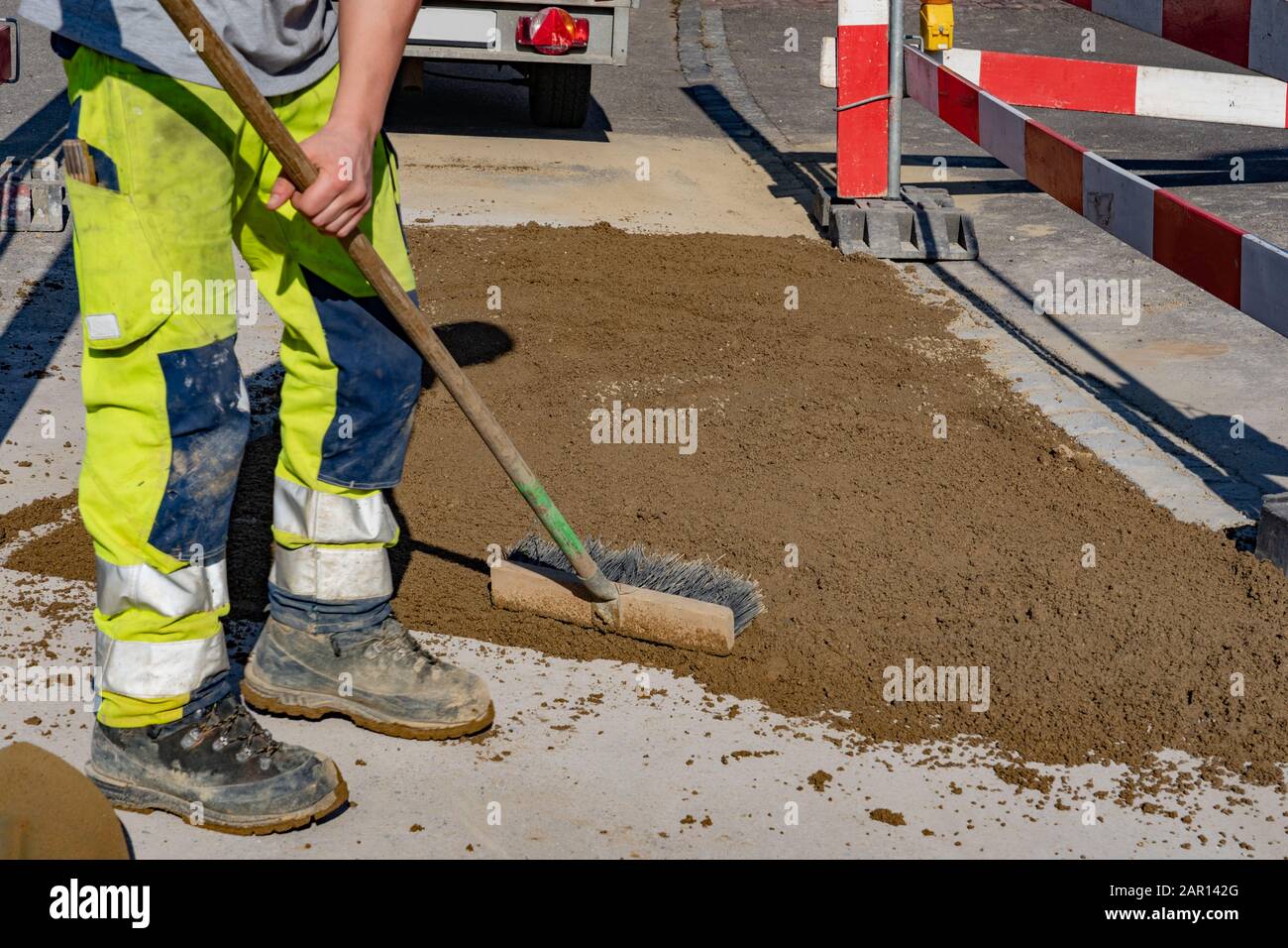 Worker with warning trousers and working shoes holding a street broom in his hand and standing near sand area on the street. Sand scoop; shovel; barri Stock Photo