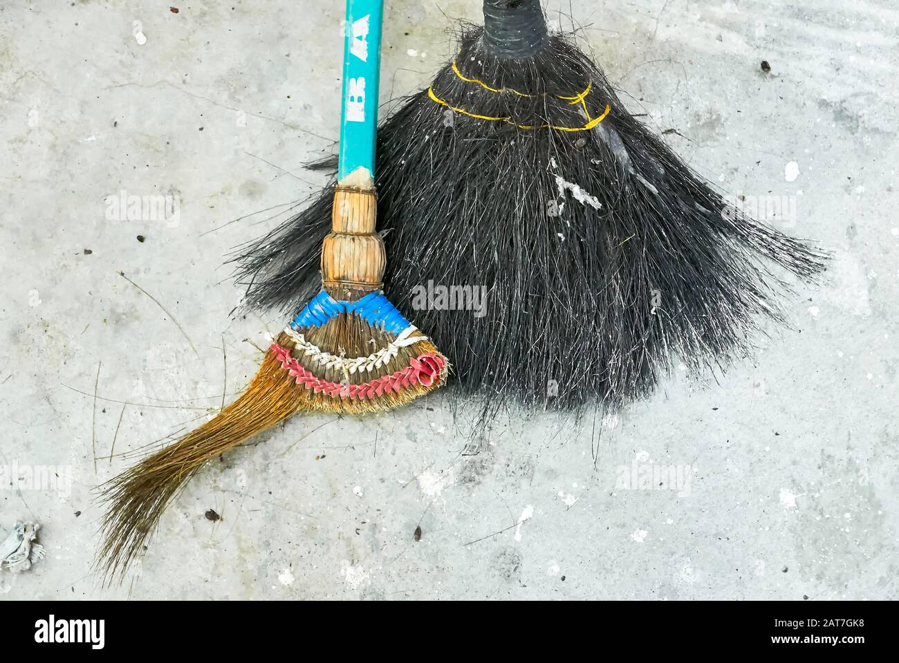 Close-up of two overused old broken brooms standing on a grey concrete floor next to each other, seen in the Philippines Stock Photo