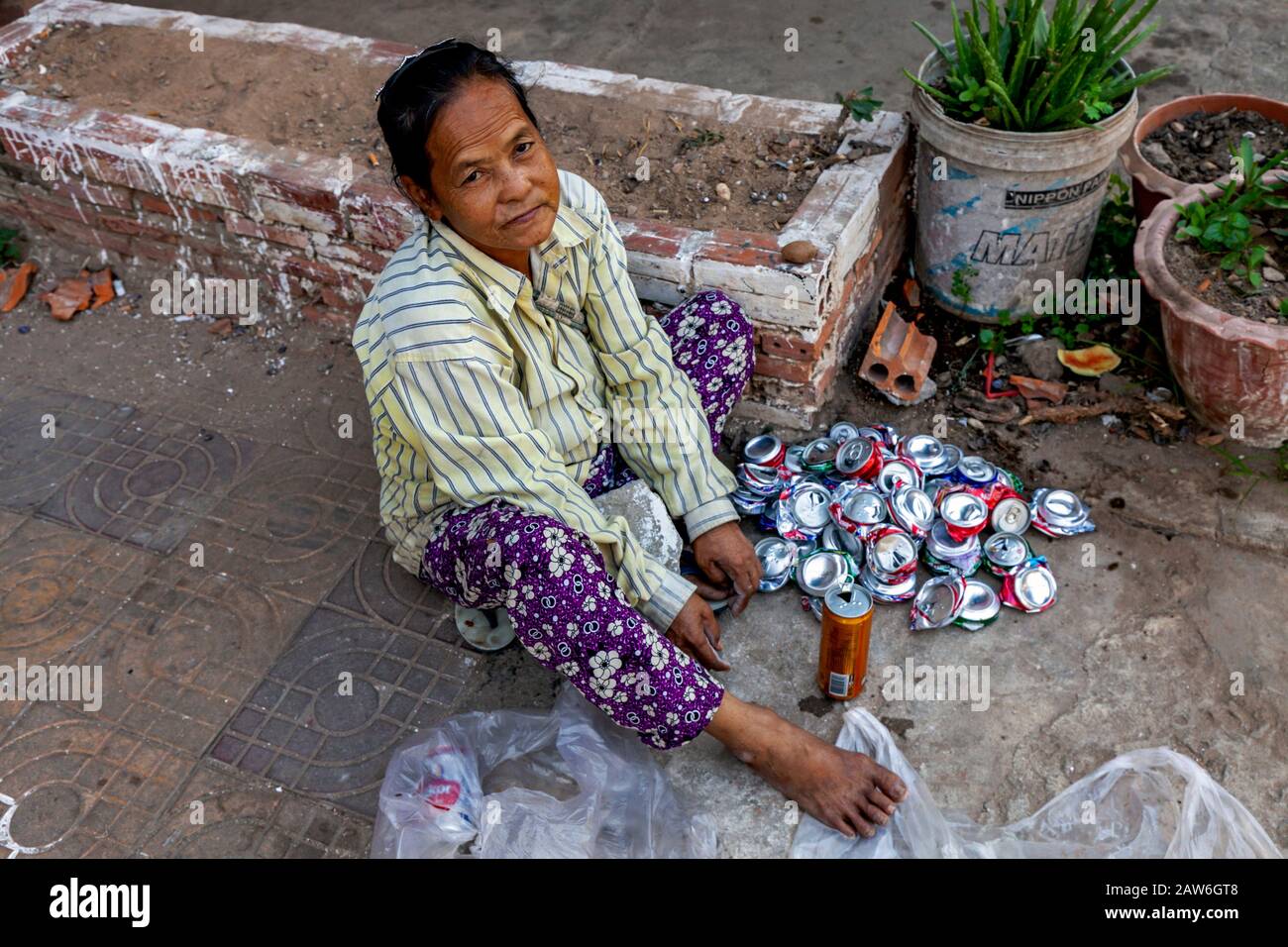 A 50 year old Cambodian woman living in poverty sits on a sidewalk crushing recyclable aluminum cans in urban Kampong Cham, Cambodia. Stock Photo