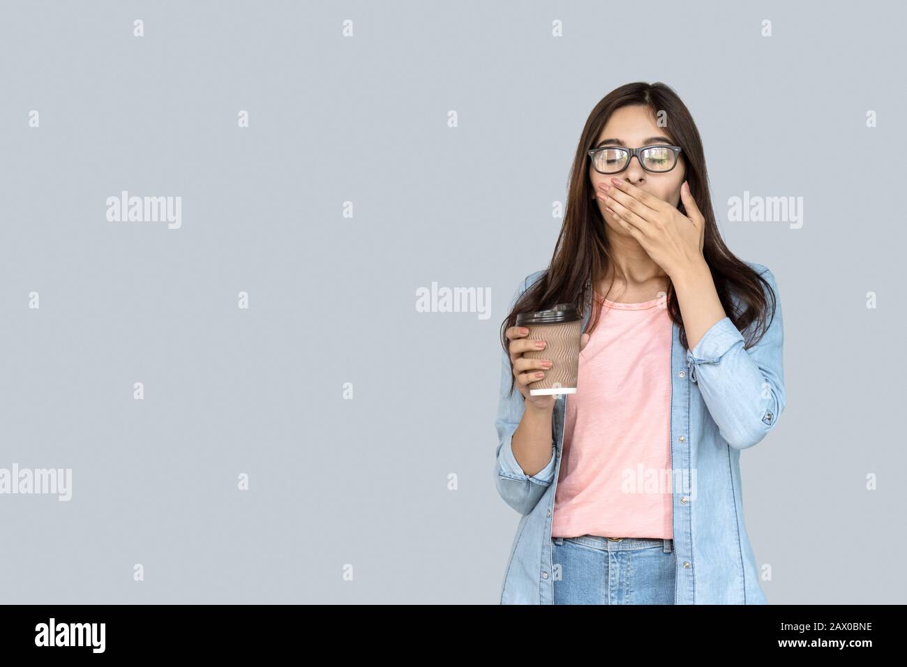 Young tired sleepy indian girl yawning holding coffee isolated on gray Stock Photo