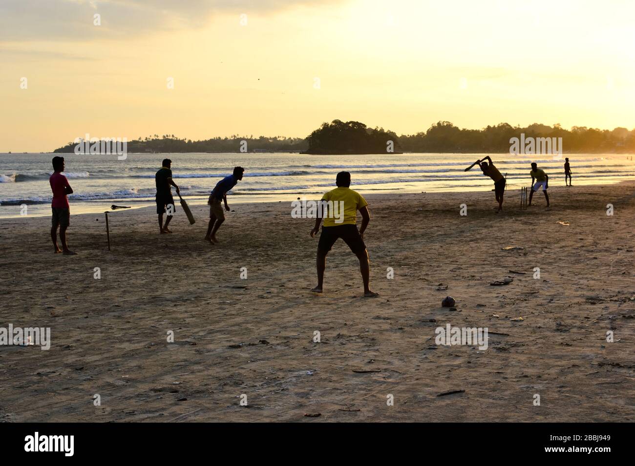 Young boys playing cricket game on a beach. Silhouettes of young  boys having fun on Sri Lanka beach. Golden sunset light with sunbeams in Weligama Stock Photo