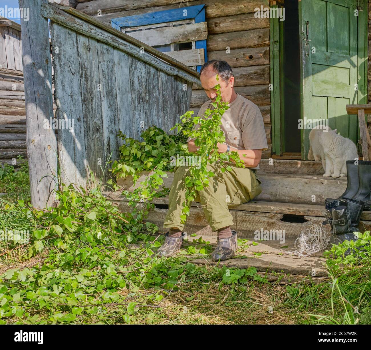 Senior Asian man ties birch brooms for a bath sitting on a wooden porch of an old country house in a Siberian village, Russia Stock Photo