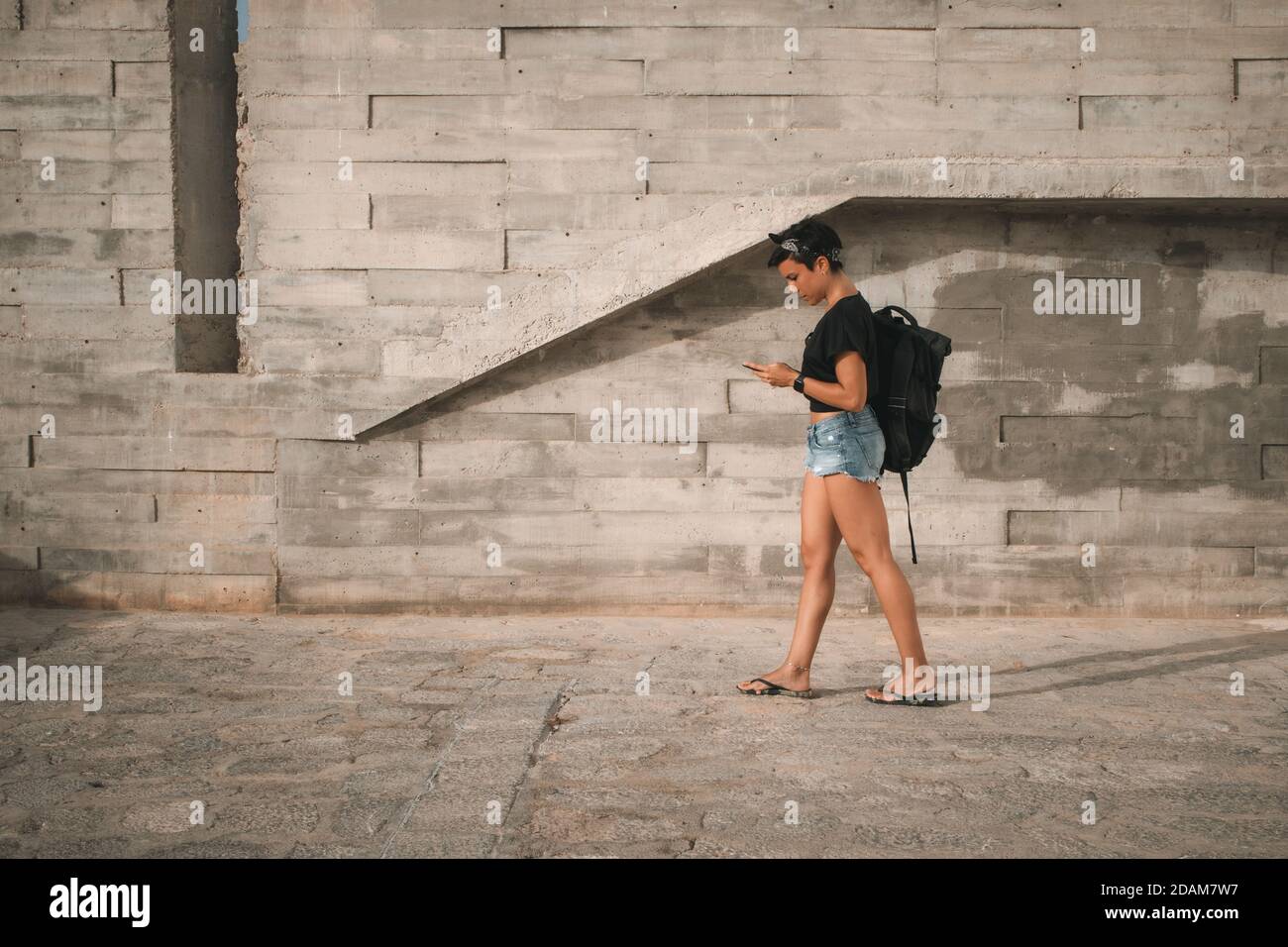 Attractive woman with short hair wearing summer clothes and a backpack walking while looking at her phone and texting Stock Photo