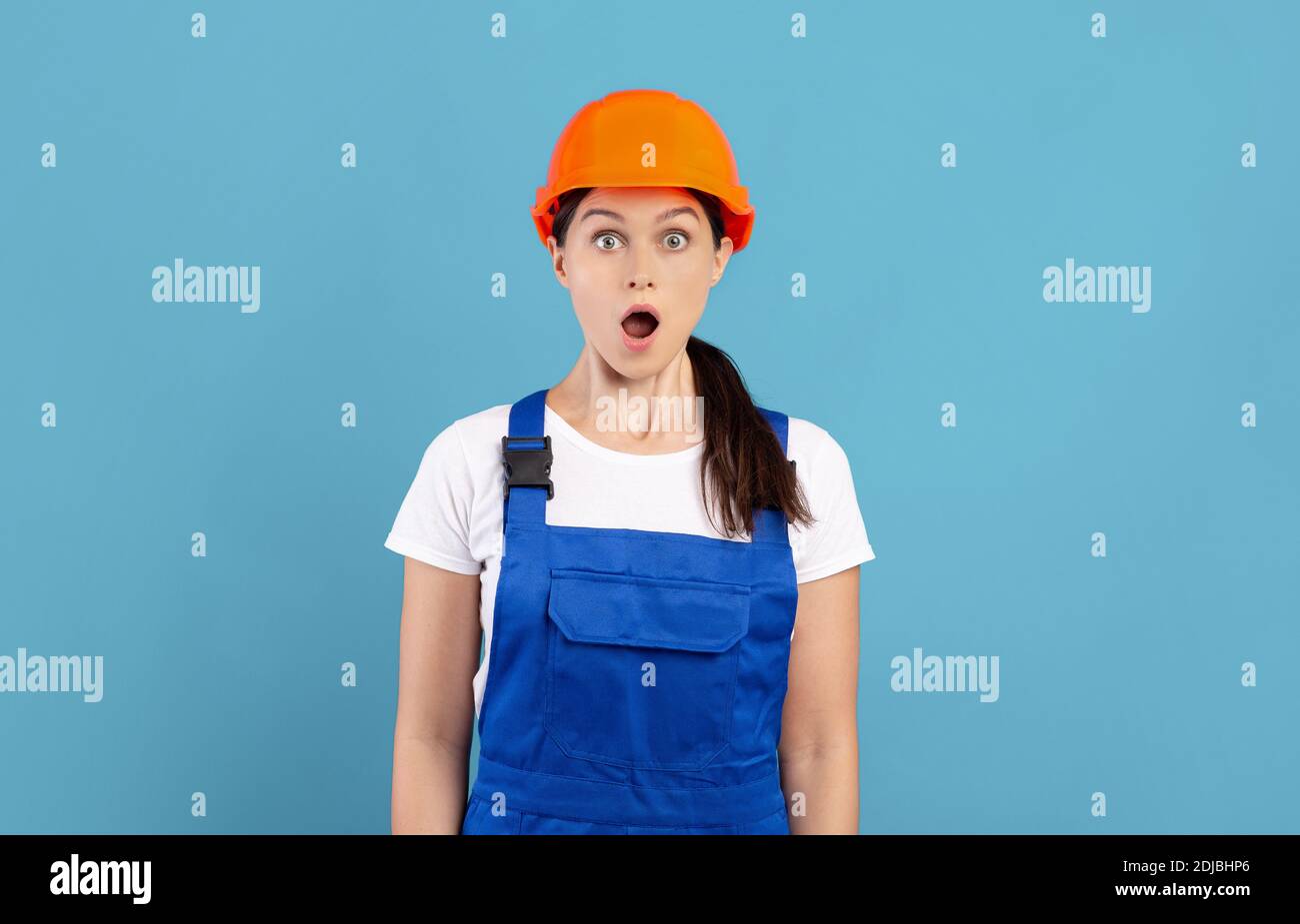 Portrait Of Shocked Handywoman In Hardhat And Coveralls Standing Over Blue Background Stock Photo