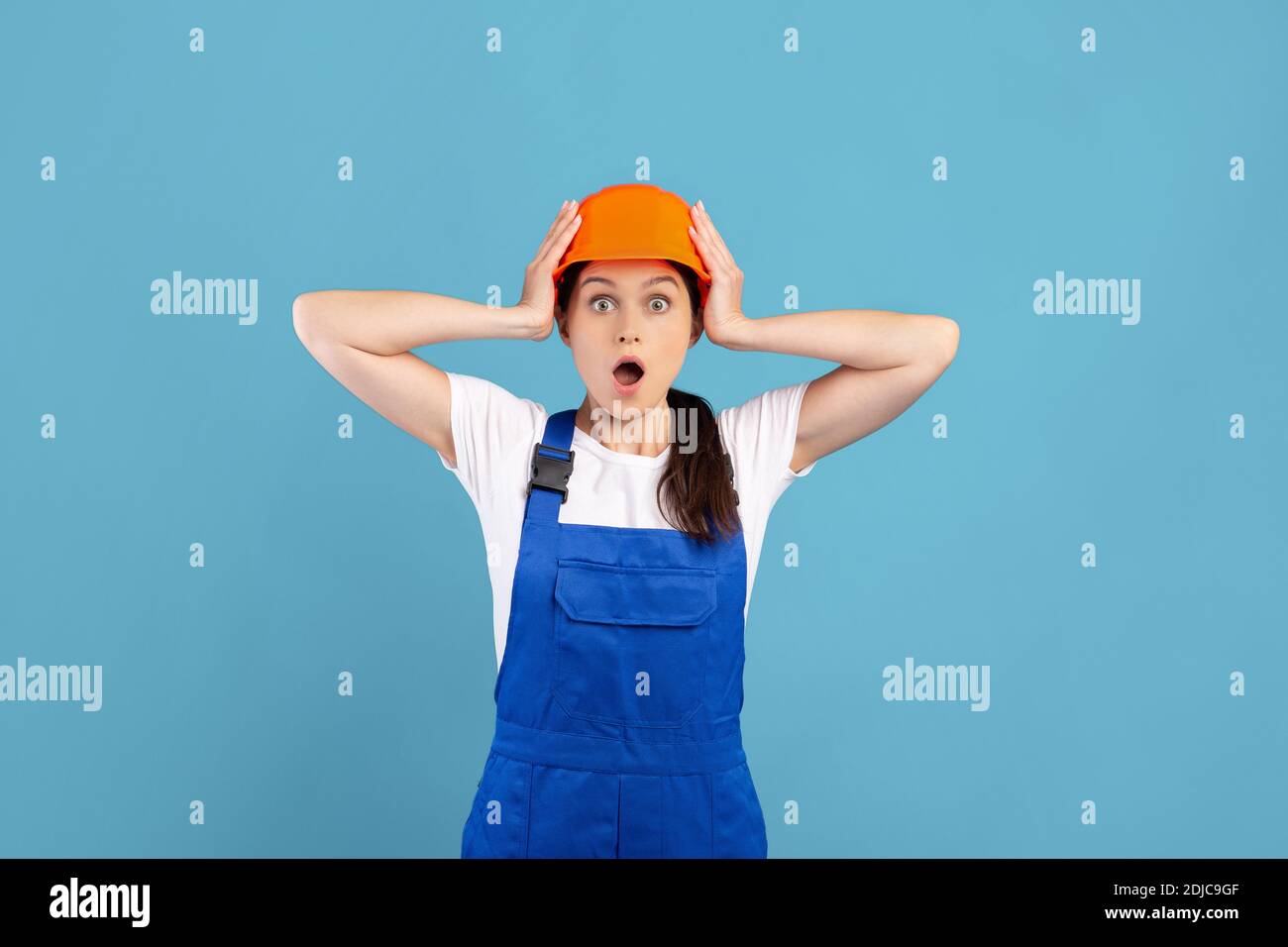 Shocked handywoman in protective helmet and coveralls touching head in excitement Stock Photo