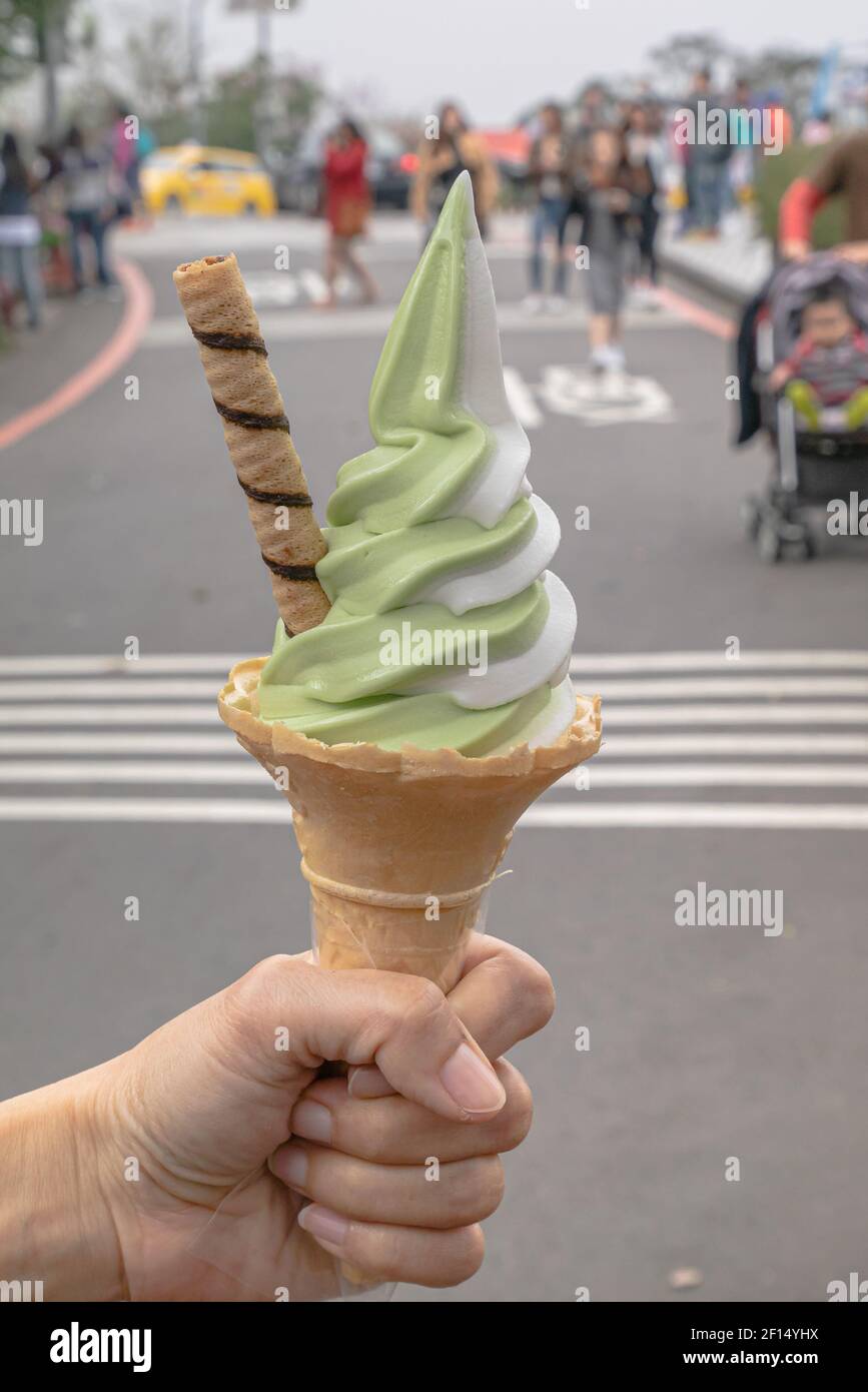 Ice cream cone, a close up of matcha green tea and vanilla soft-served with waffle cone in hand holding at travel street with people background in Tai Stock Photo