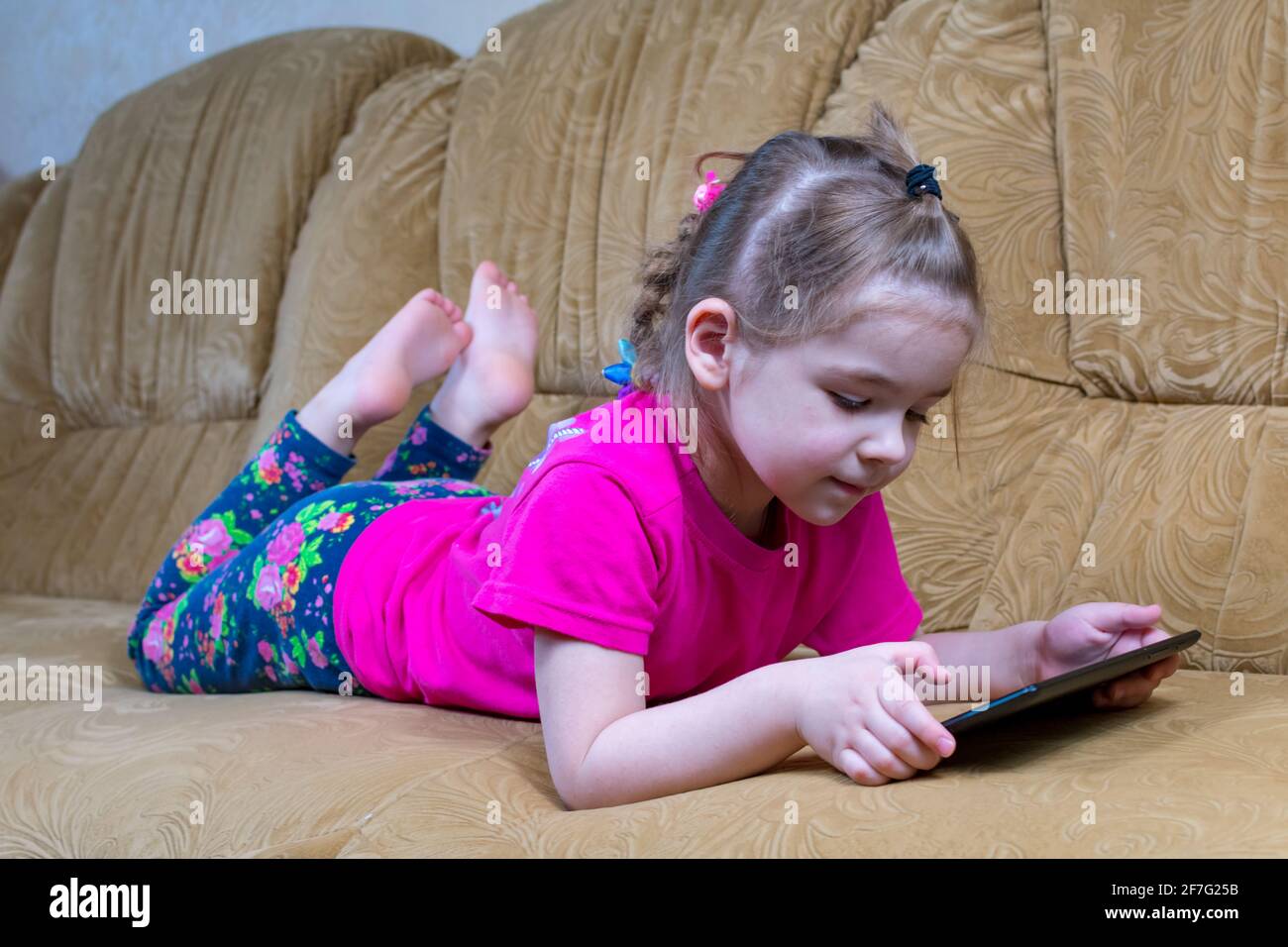 Little girl lying on a comfortable sofa enjoying an online game on a digital tablet computer. Addicted to technology, happy little kid using fun apps Stock Photo