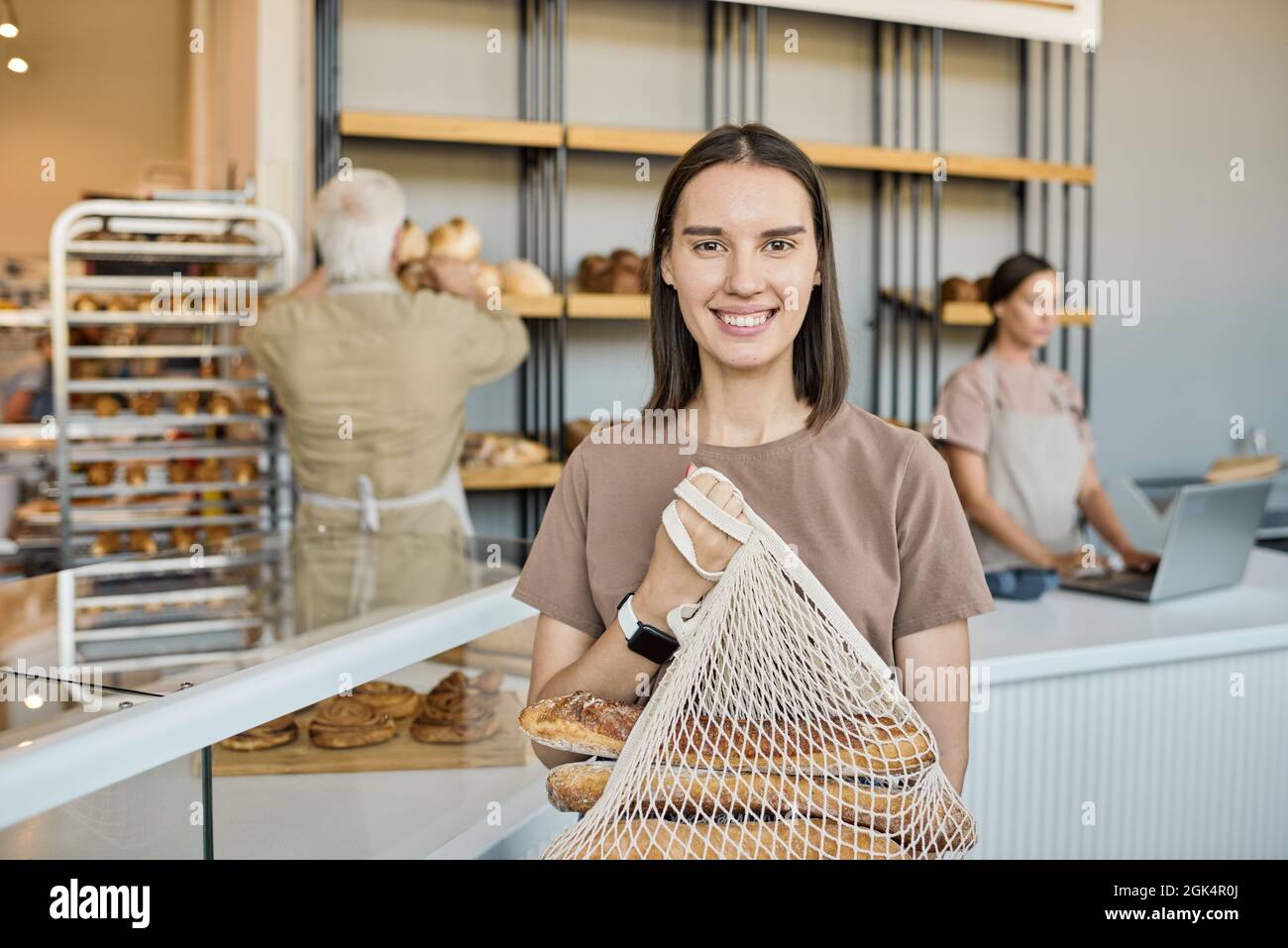 Portrait of happy young female customer holding net bag with fresh loaves of bread against bakery workers Stock Photo