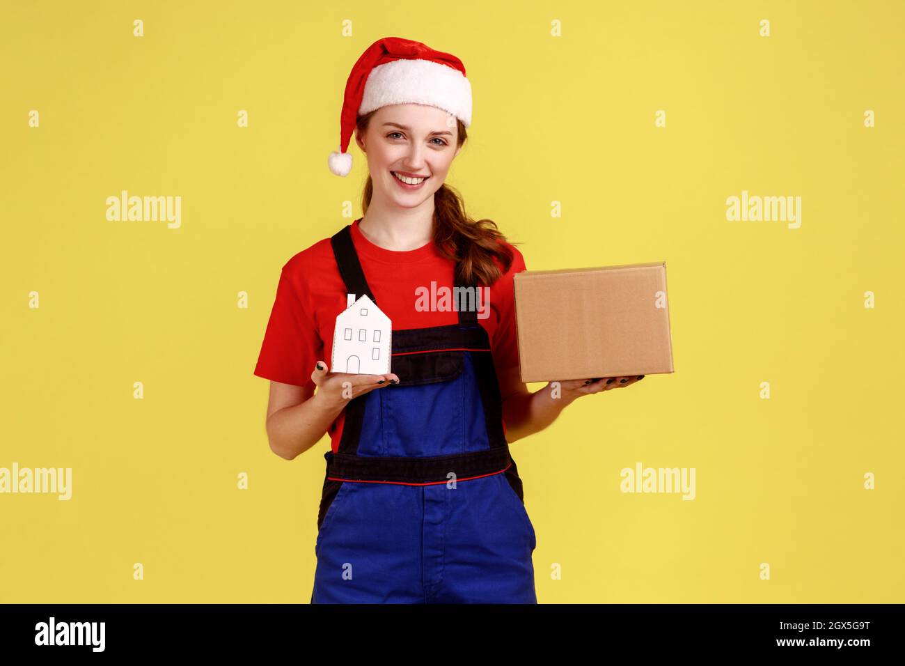 Smiling woman mover or worker holding cardboard parcel and paper house, relocation service, wearing blue overalls and santa claus hat. Indoor studio shot isolated on yellow background. Stock Photo