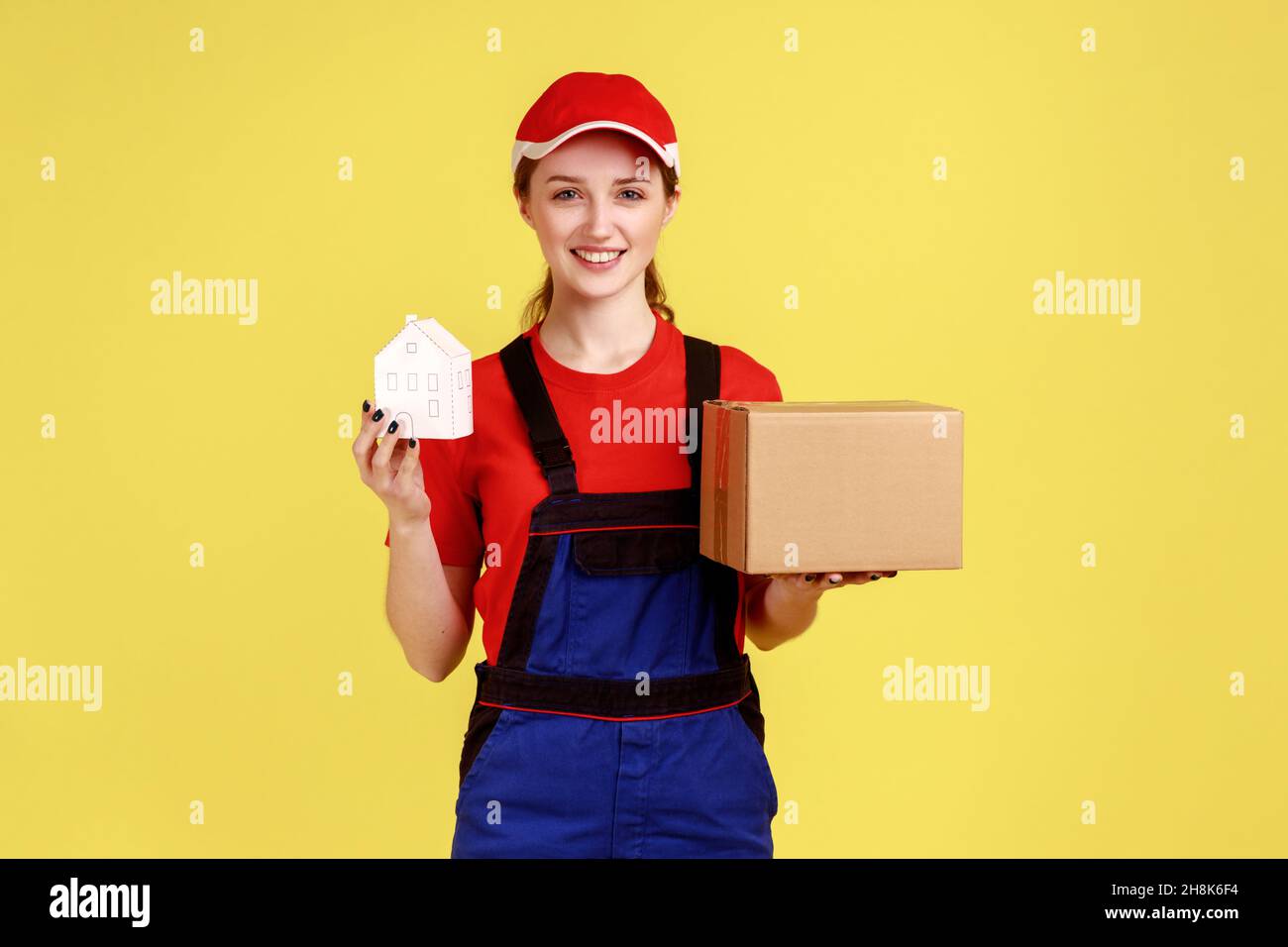 Satisfied beautiful courier woman holding parcel and paper house in hands, fast delivery door-to-door, wearing work uniform and red cap. Indoor studio shot isolated on yellow background. Stock Photo