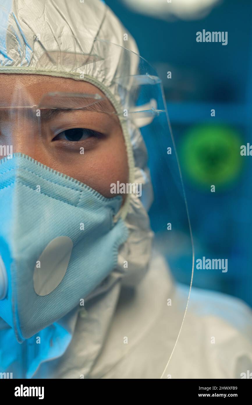 Half of face of young Asian woman in coveralls, respirator and protective eyeglasses standing in front of camera in scintific laboratory Stock Photo