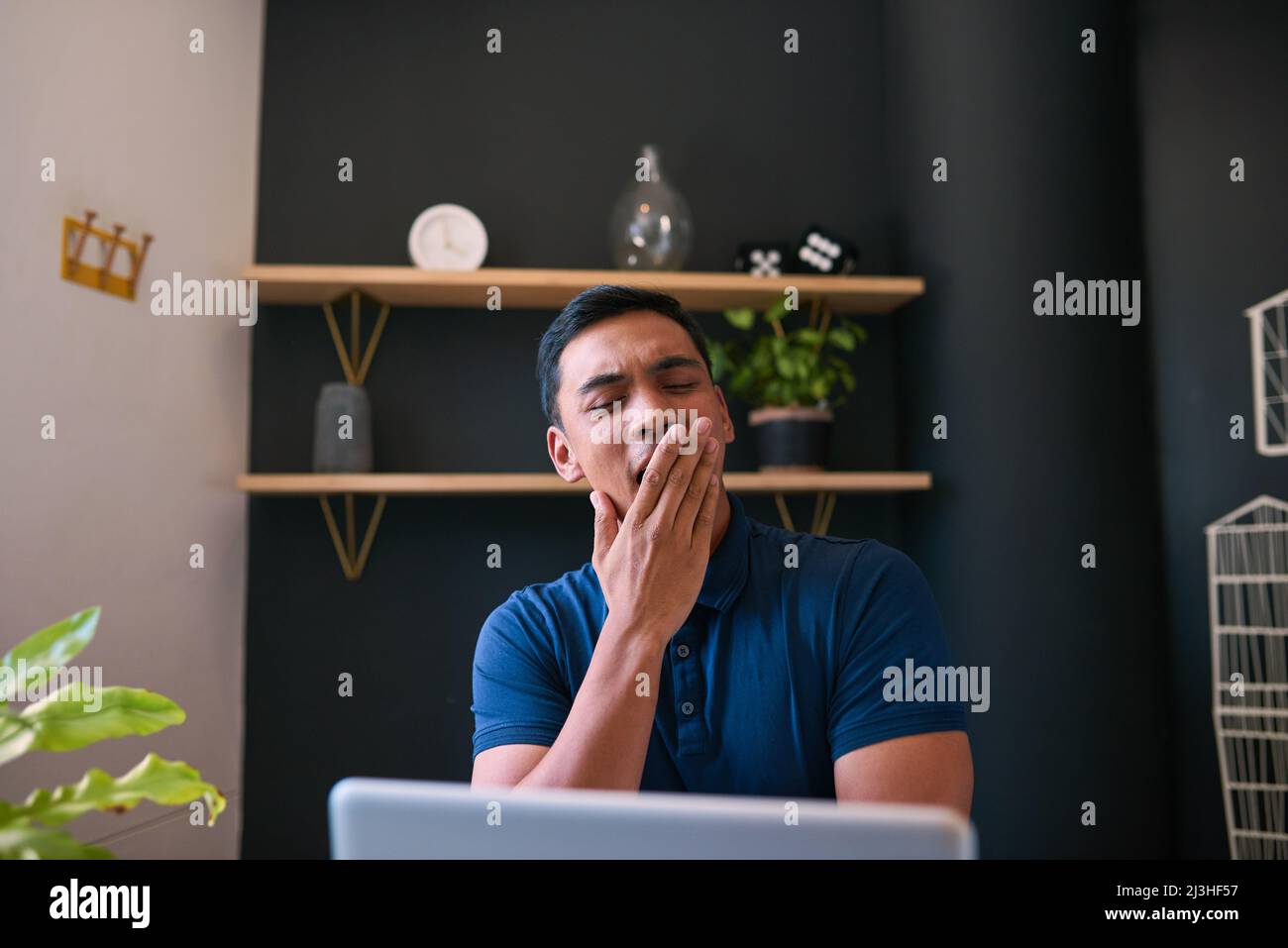 A young Asian man yawns at his desk in the office with his hand over mouth Stock Photo