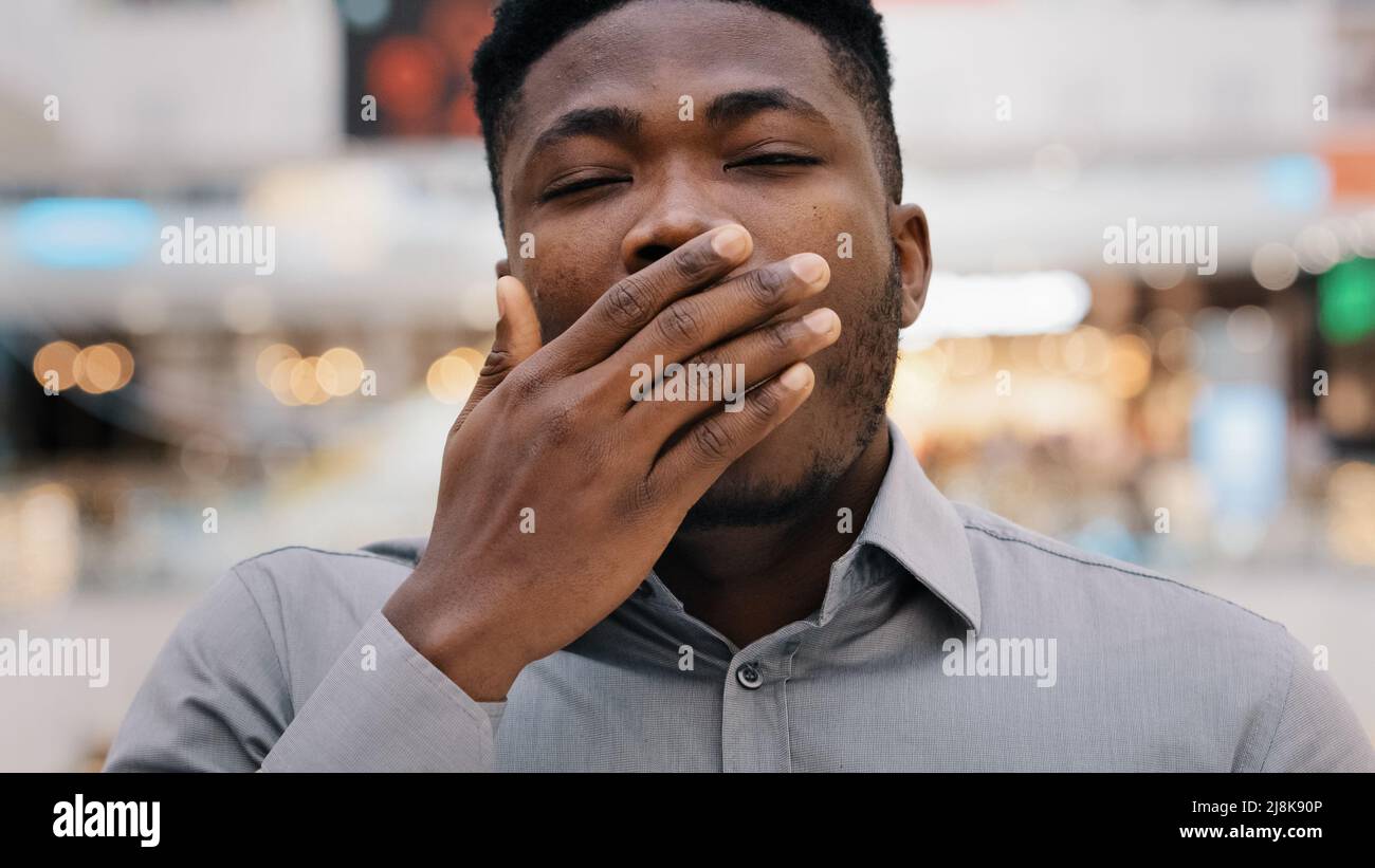 Male portrait close-up man overworked young african american man tired worker looking at camera yawns covers mouth with hand bored feels tired drowsy Stock Photo