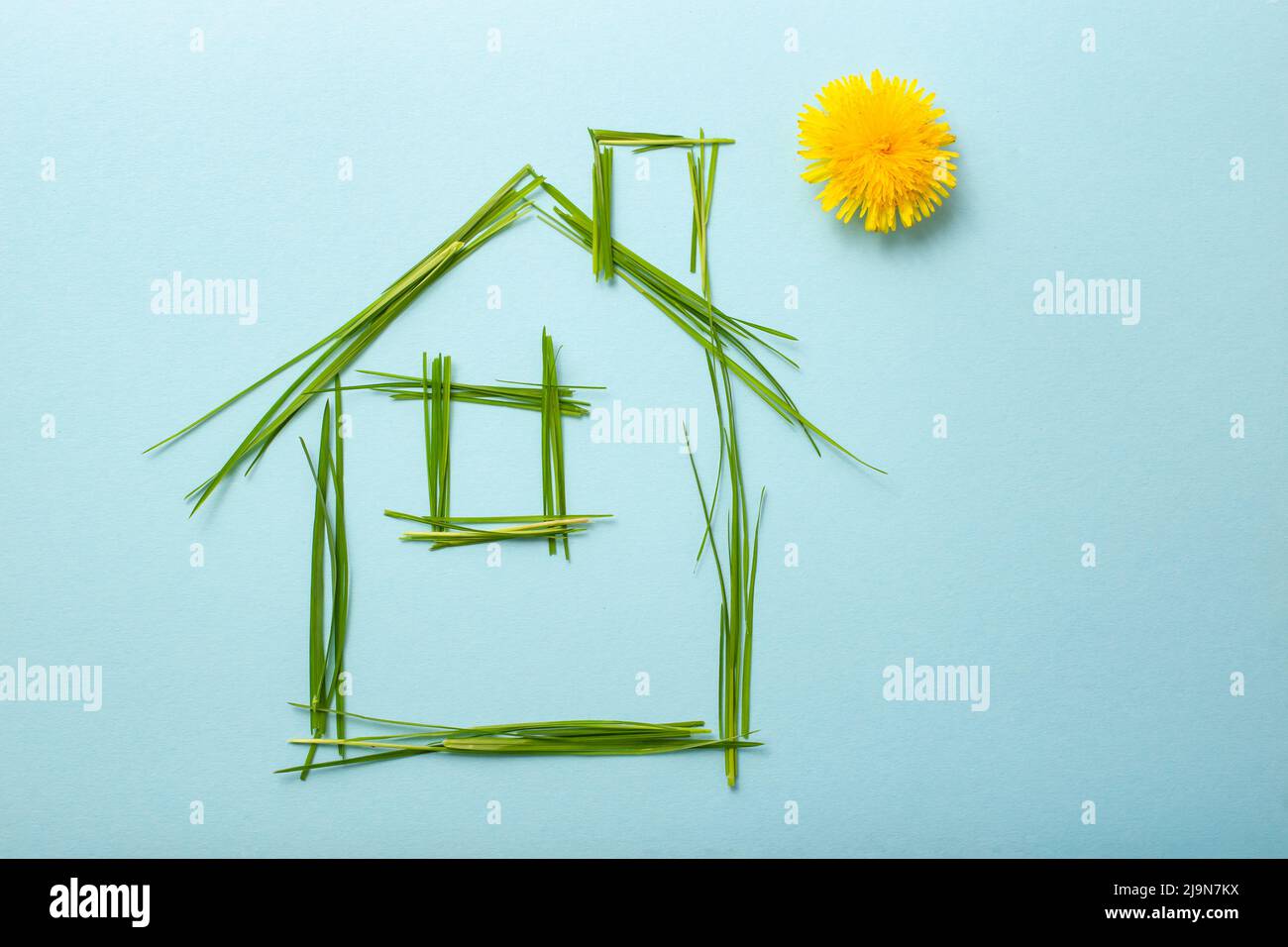 house made of grass bades with chimney and a dandelion as the sun. Grass straws on light blue background Stock Photo