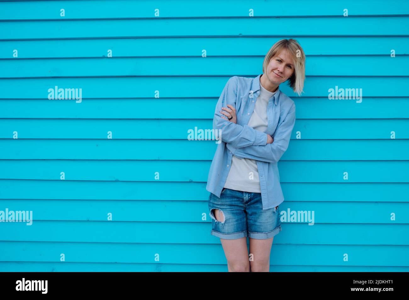 Middle-aged woman wearing grey T-shirt, blue shirt, shorts, leaning against blue wall with crossed arms, bowing head. Stock Photo