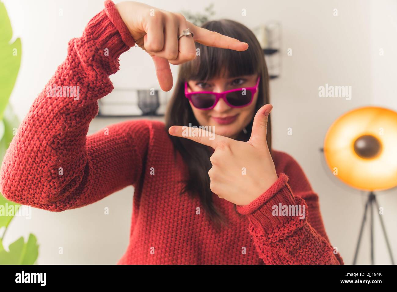 Caucasian woman with dark hair wearing pink sunglasses and red sweater framing face with hands. Indoor shot. High quality photo Stock Photo