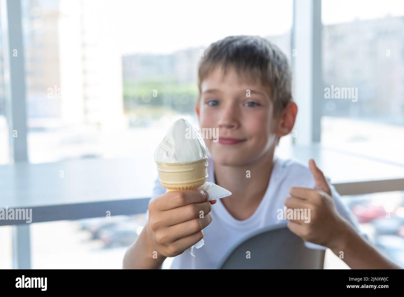 A cute boy eats white ice cream in a waffle cup and shows a thumbs-up gesture while sitting at a table by the window in a cafe. Blurred background. A Stock Photo