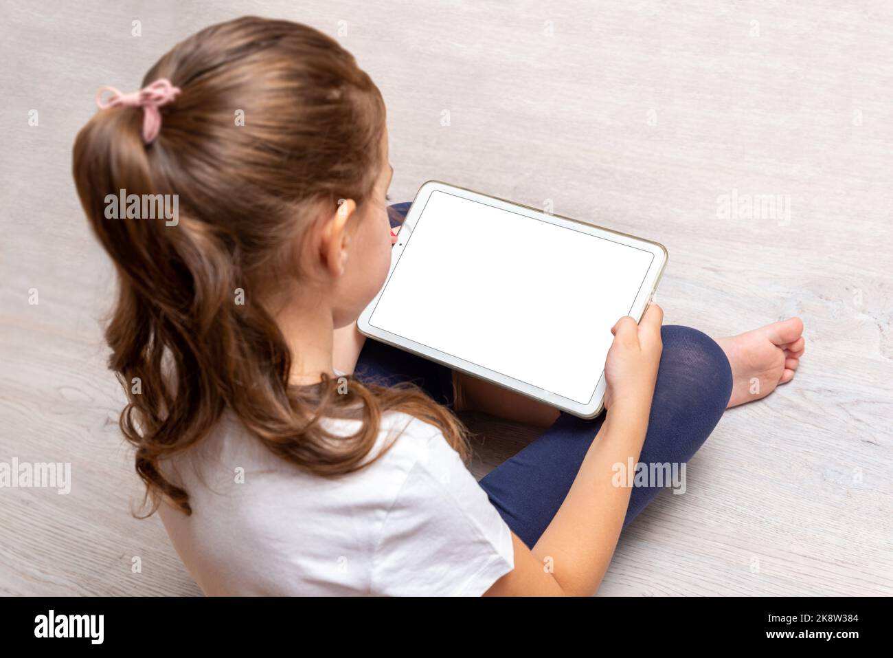 Little girl sitting on the floor and holding a white tablet with isolated screen for promoting video games, websites or apps Stock Photo