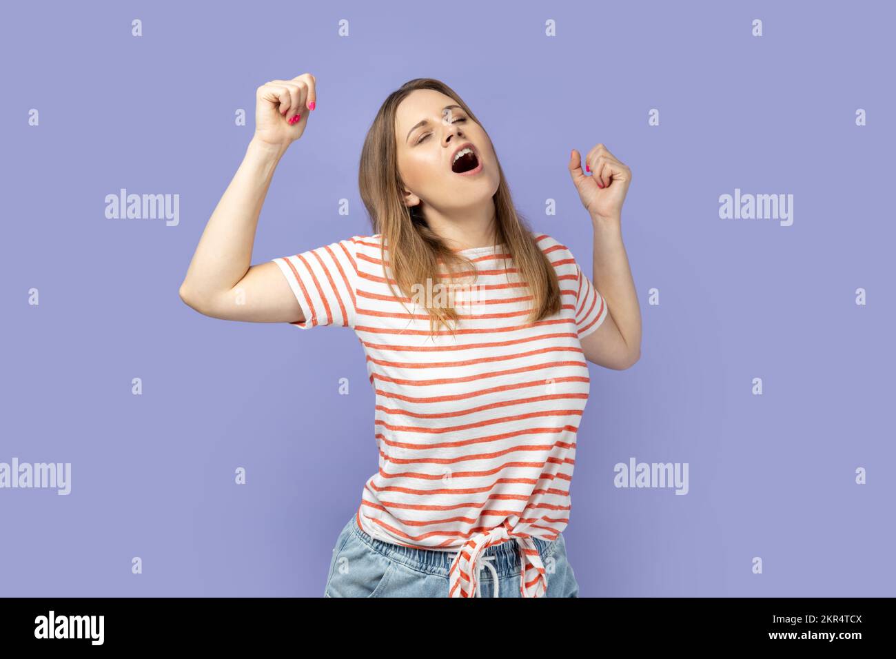 Portrait of tired blond woman wearing striped T-shirt standing yawning with closed eyes, stretching arms, feeling exhausted need rest. Indoor studio shot isolated on purple background. Stock Photo