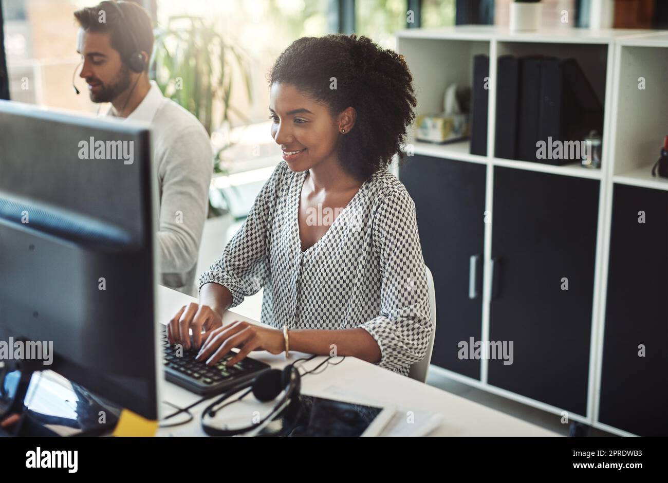 Her positive attitude is infectious. a young businesswoman working on her computer with her colleague in the background. Stock Photo