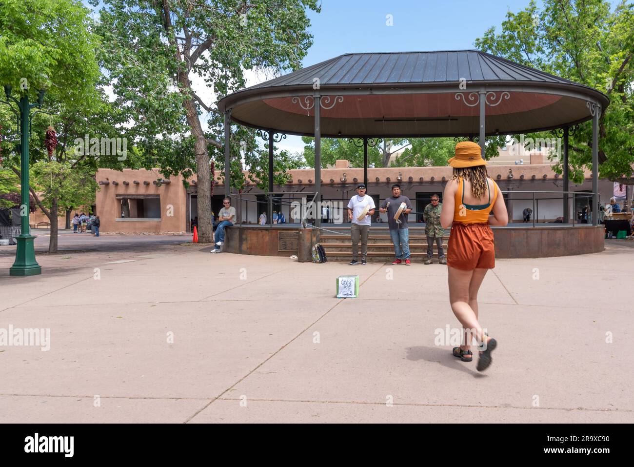 A young woman wearing a modish orange color coordinated short shots, sleeveless top and hat walks to put money in buskers',  collection box Stock Photo