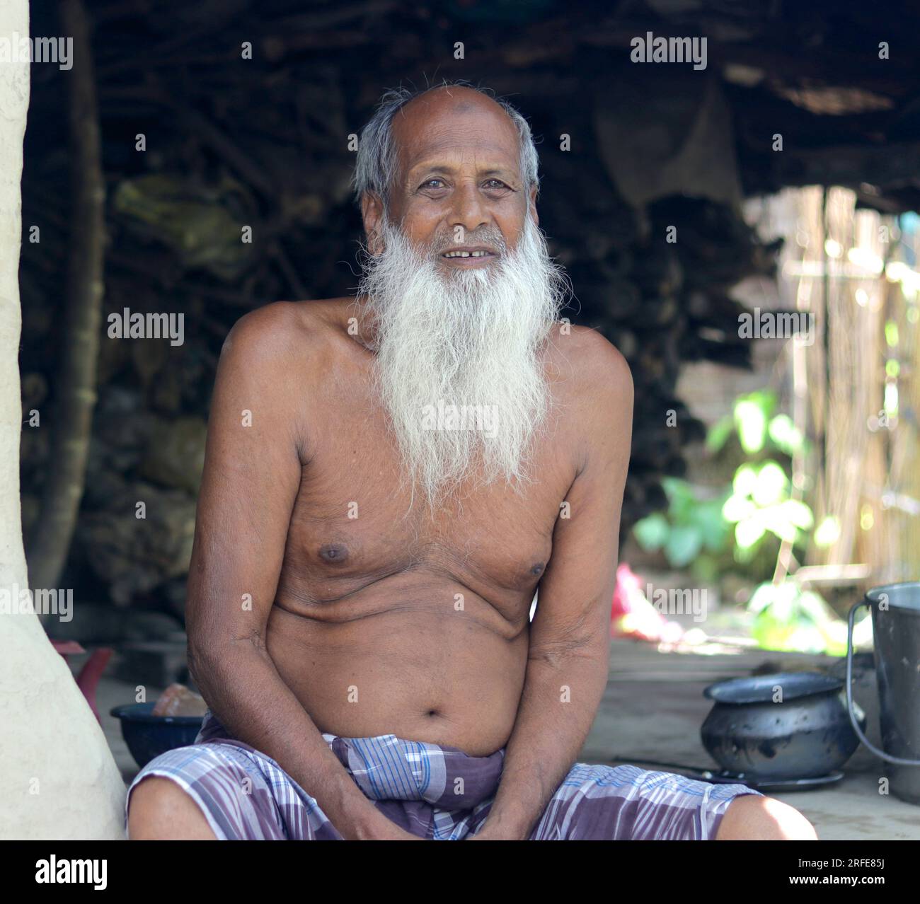 An old poor man with a long white beard sitting on his village mud house. Stock Photo