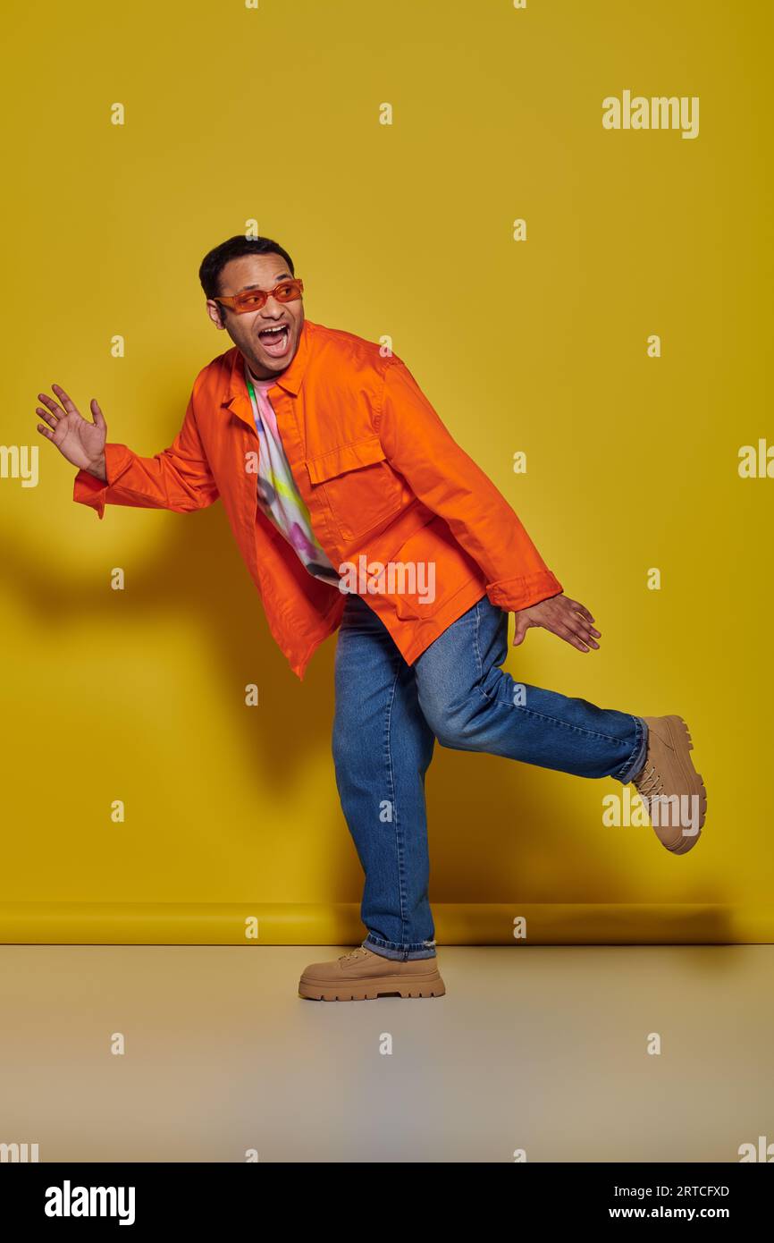 scared indian man in sunglasses and vibrant attire running away and screaming on yellow backdrop Stock Photo