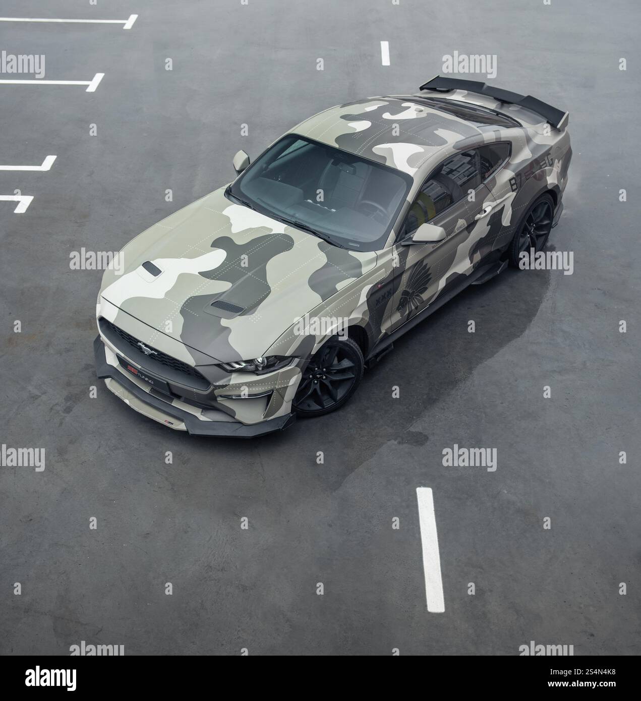 Green camo Ford Mustang in parking lot. Aerial three quarter front view of american pony car parked on wet concrete surface. Stock Photo