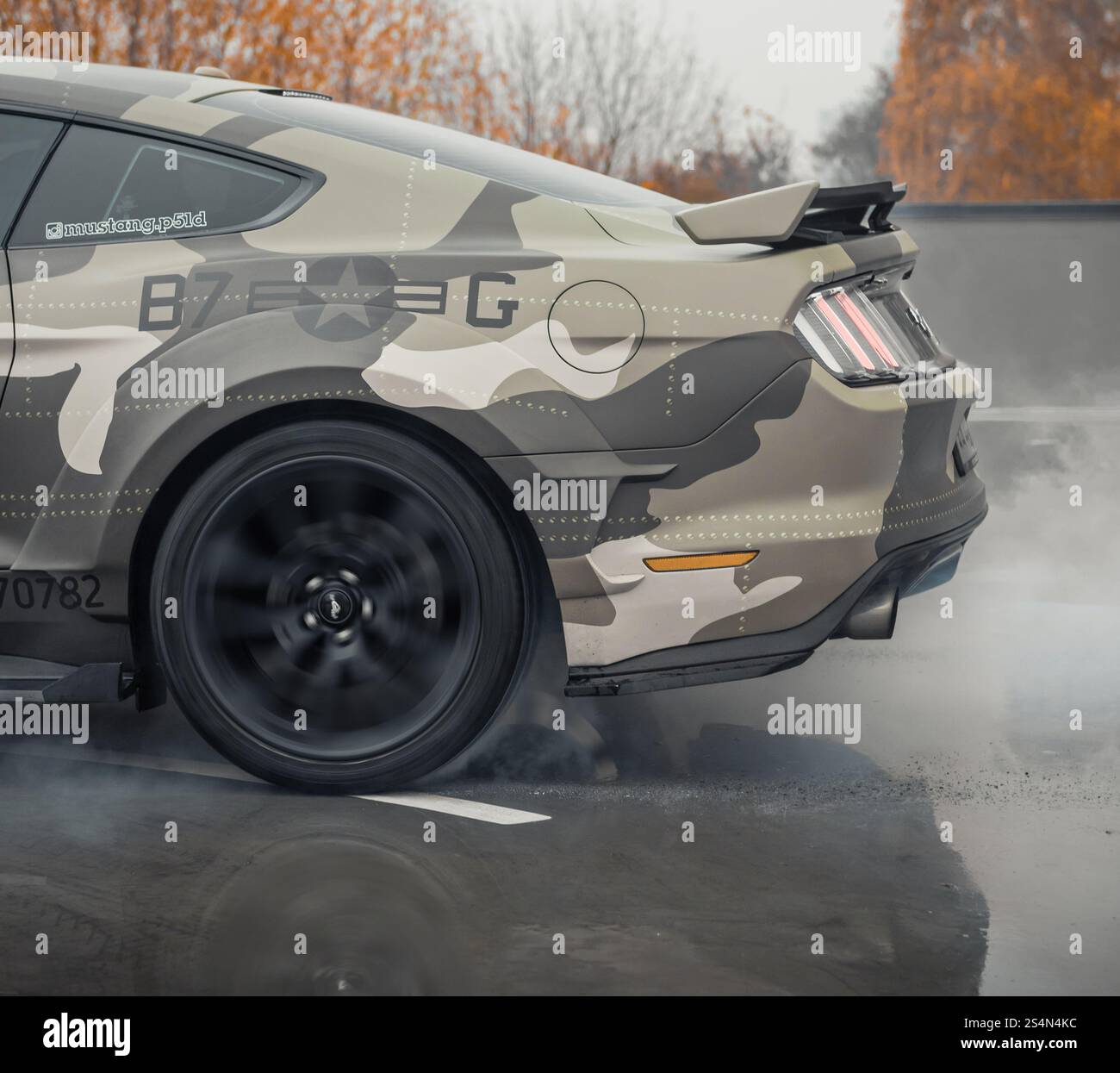 eige camo Ford Mustang doing a burnout. Rear side view of american pony car burning rubber on wet concrete surface. Stock Photo