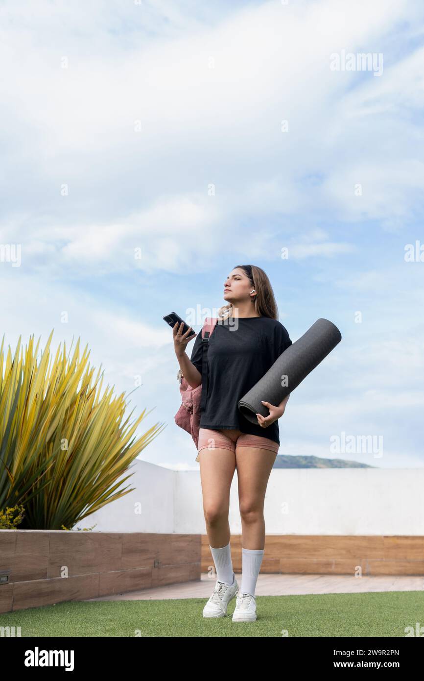 Young Latina woman wearing shorts and black t-shirt walking with a matt and her cell phone towards the gym Stock Photo