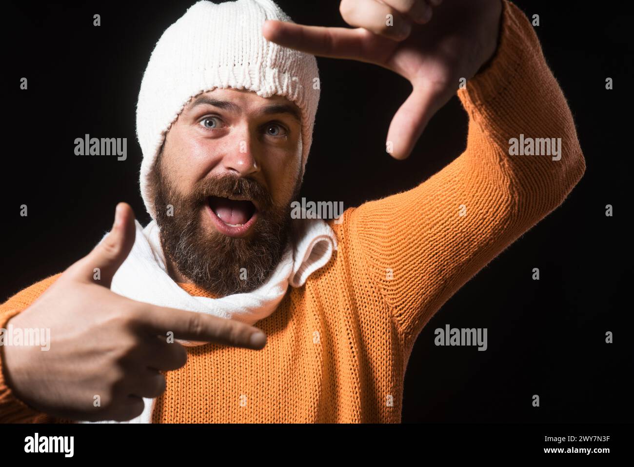 Happy man in orange sweater, white hat and scarf makes camera frame with fingers. Bearded man using hand framing to create viewfinder. Imaginary Stock Photo