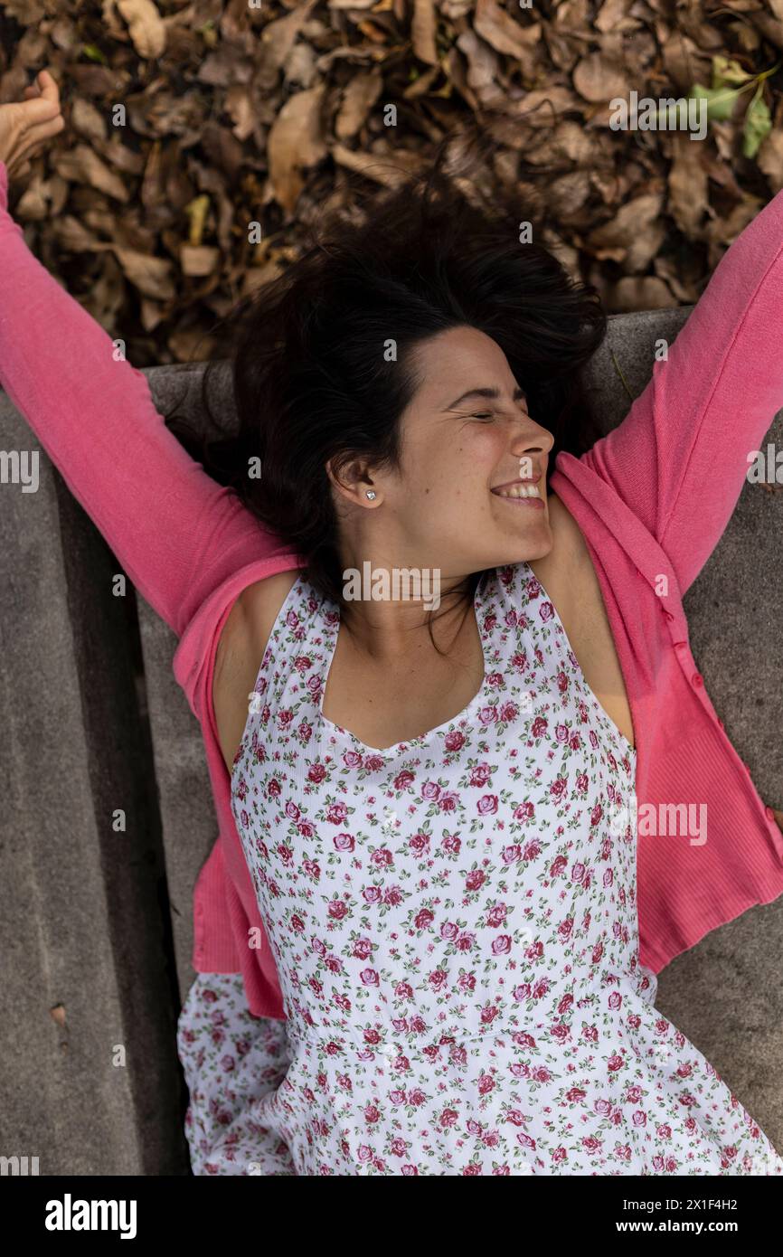 Young Latin American woman (33) with positive attitude is very happy for the arrival of spring. She is lying on her back, background of dry leaves. Sh Stock Photo