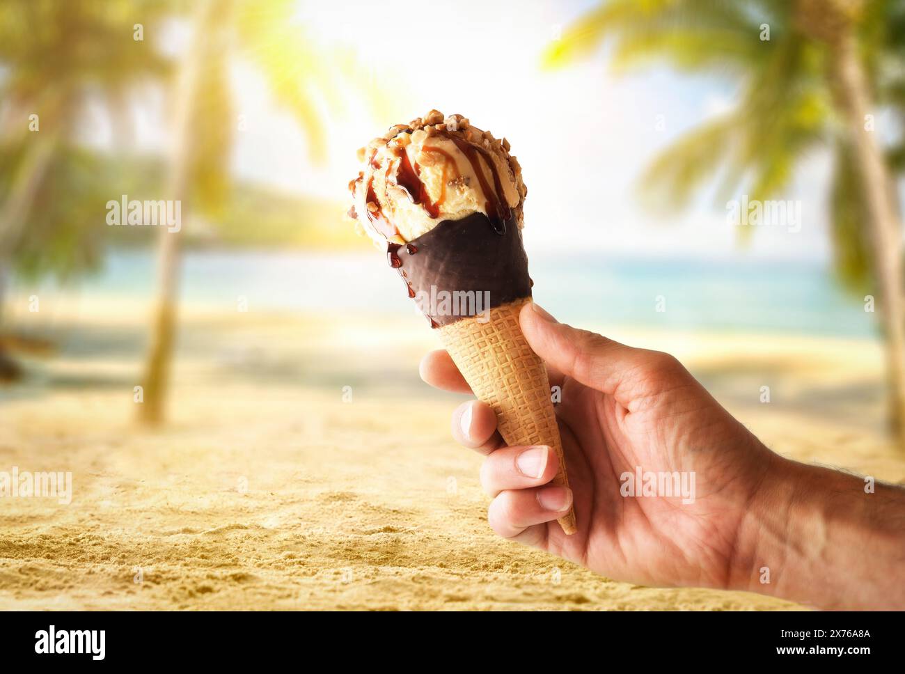 Hand holding a chocolate ice cream cone with vanilla scoop with caramel and almond pieces on vacation with beach and palm trees background. Stock Photo