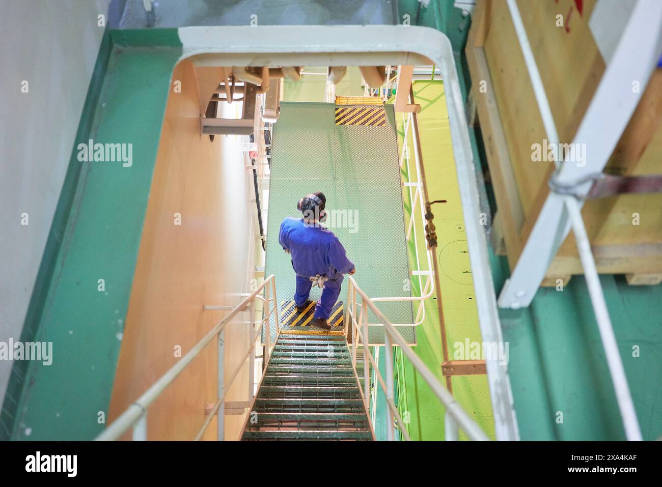 A person in blue coveralls is descending a metal staircase in an industrial environment with yellow safety rails. Stock Photo