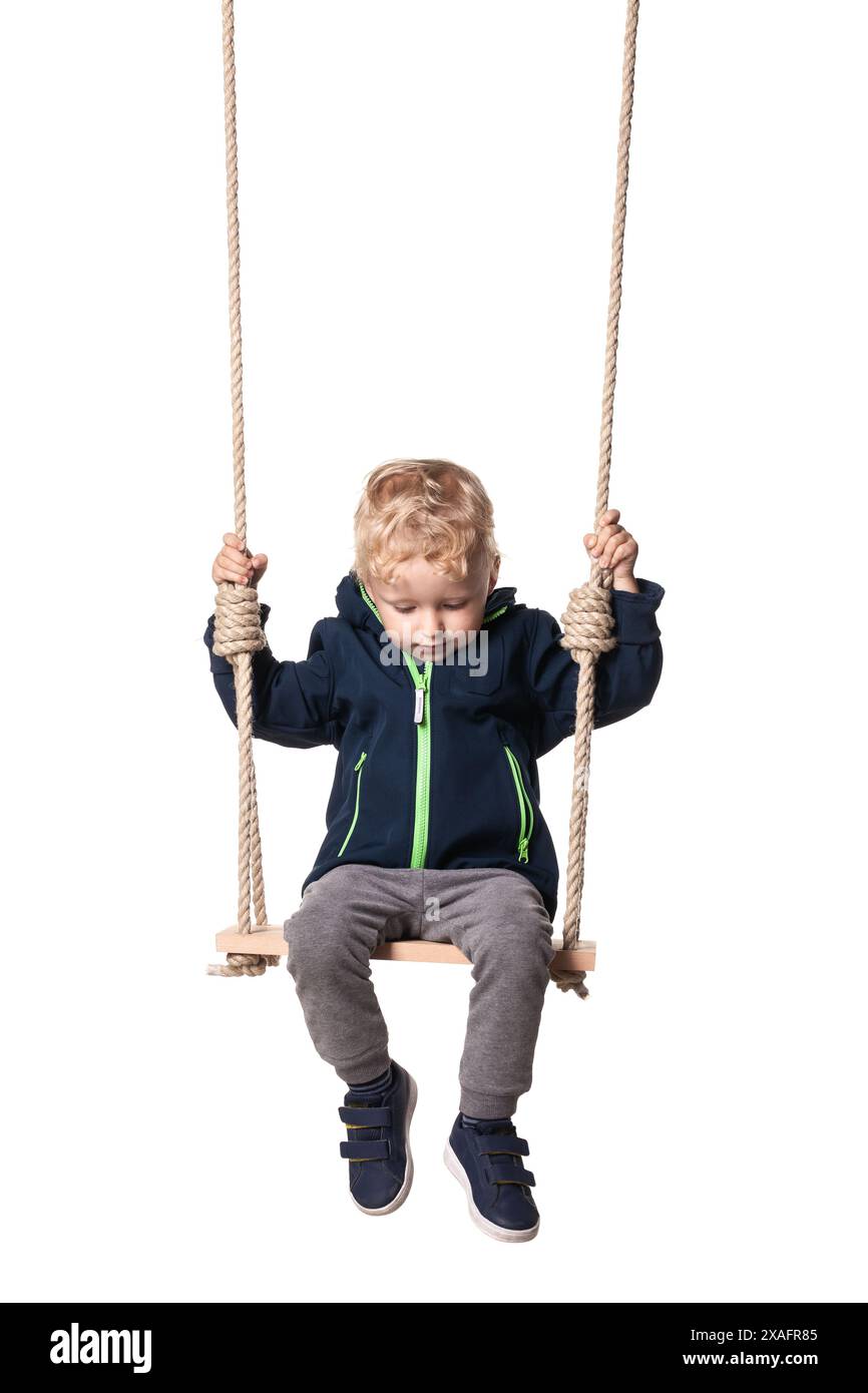 Cute little boy in blue jacket and grey pants sitting on a swing, isolated on white background. Stock Photo