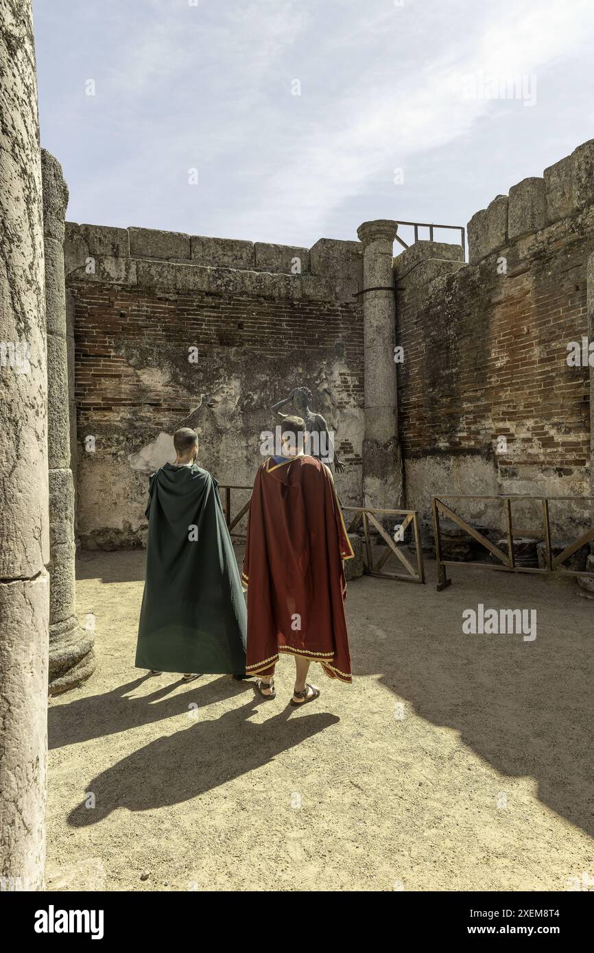 Two men dressed in Roman-style cloaks observe a statue at an ancient Roman archaeological site in Merida, Spain, surrounded by historic stone columns Stock Photo