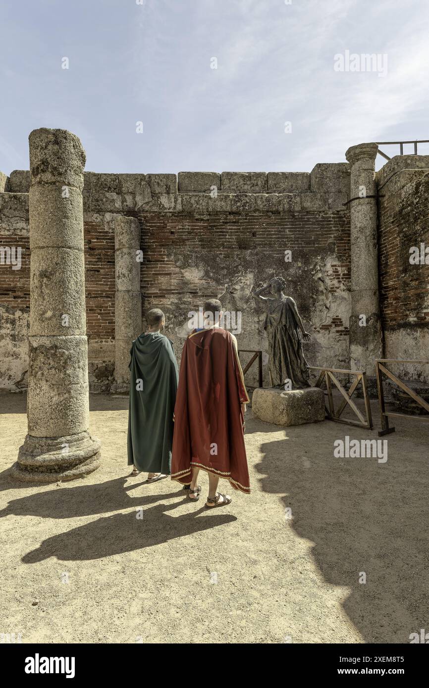 Two men dressed in Roman-style cloaks observe a statue at an ancient Roman archaeological site in Merida, Spain, surrounded by historic stone columns Stock Photo