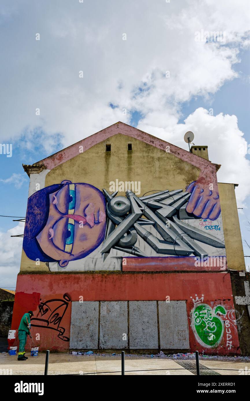 A painter in green starts to paint over graffiti on a weathered building in Lisbon, under a bright blue sky with scattered clouds Stock Photo