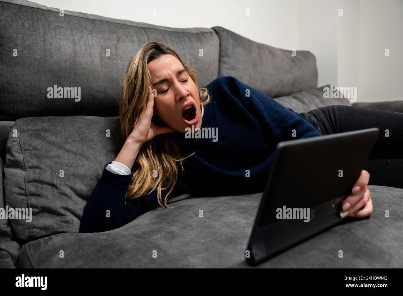 Woman lying on the sofa yawning with a tablet in her hand. Facial expression of sleep or boredom Stock Photo