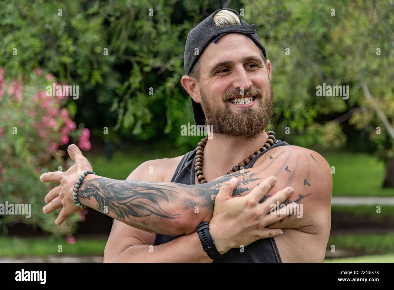 In the foreground, a young instructor smiles as he stretches his arms before beginning his outdoor class. His positive attitude and focus on wellness Stock Photo