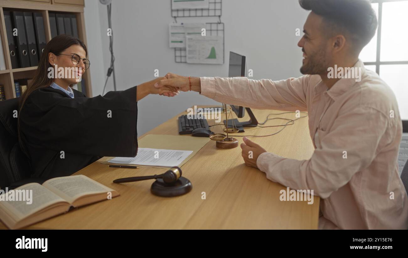 Woman judge shaking hands with man client in office setting, discussing legal matters with documents, gavel, and scales of justice on wooden desk indo Stock Photo