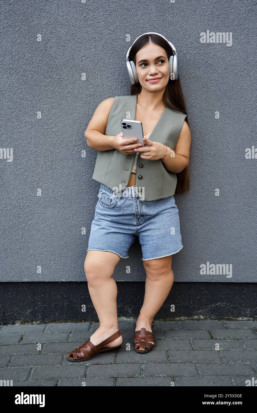 A young brunette woman with a cheerful demeanor relaxes outdoors, wearing stylish summer clothes and headphones, enjoying her music. Stock Photo