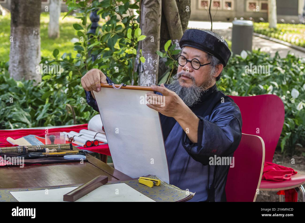 HANOI, VIETNAM, NOVEMBER 14: Old vietnamese man in national dress writes hieroglyphs on old paper at the street at November 13, 2016 in Hanoi, Vietnam Stock Photo