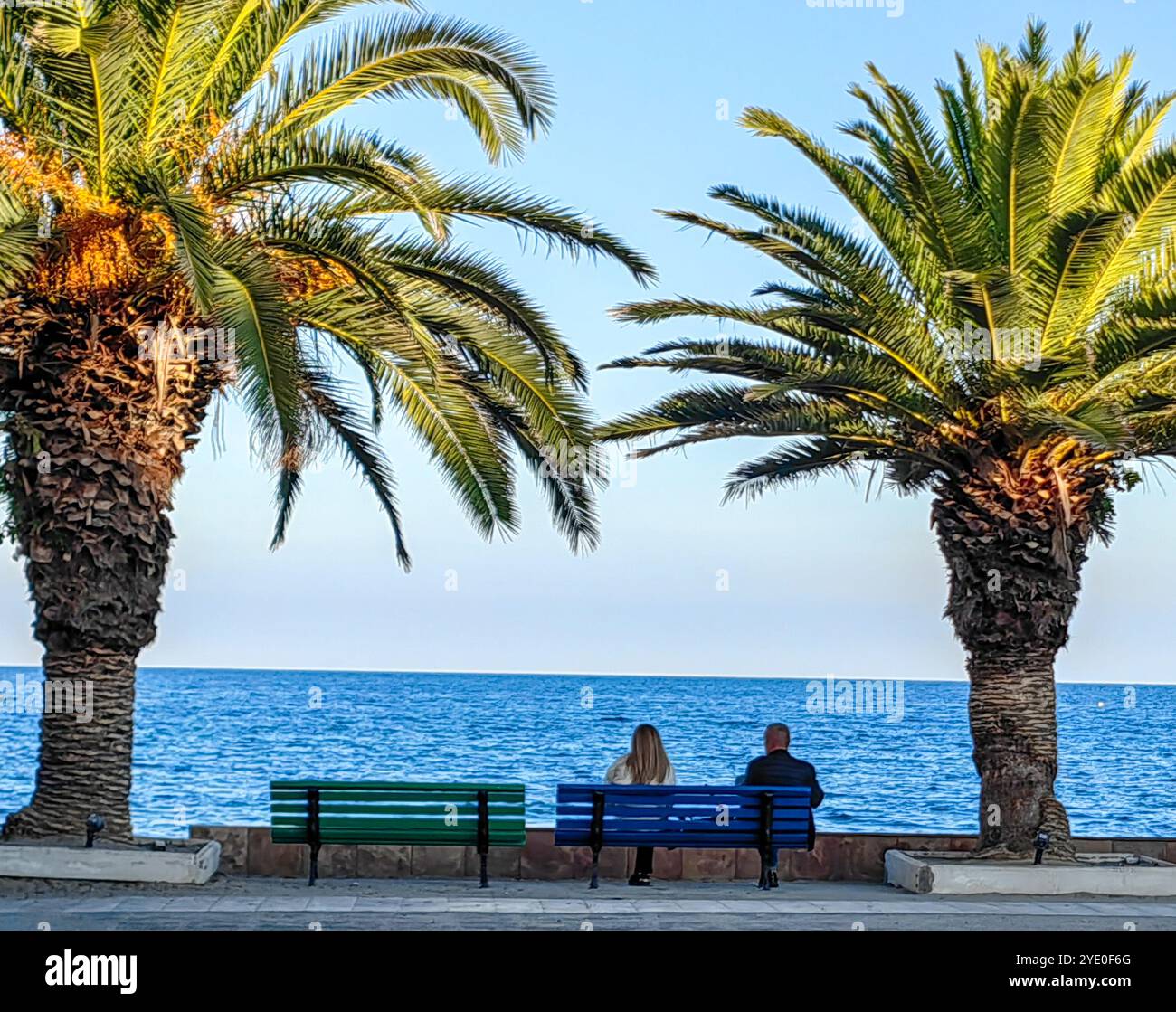 Sunny Serenity: Couple Relaxing on a Blue Bench by the Aegean Sea, Framed by Palm Trees Stock Photo