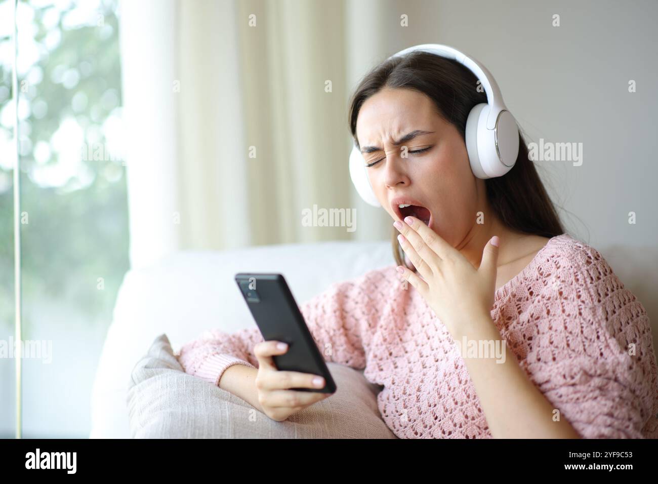 Bored woman wearing headphone yawning listening audio on phone at home Stock Photo