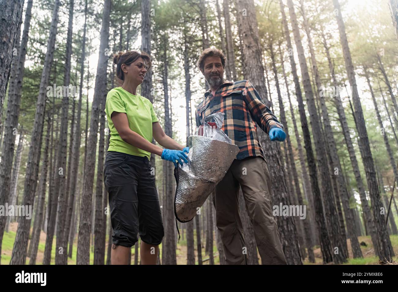 Cheerful volunteers smile while holding collected garbage in pine forest. Low angle view shows positive attitude during environmental cleanup with sun Stock Photo